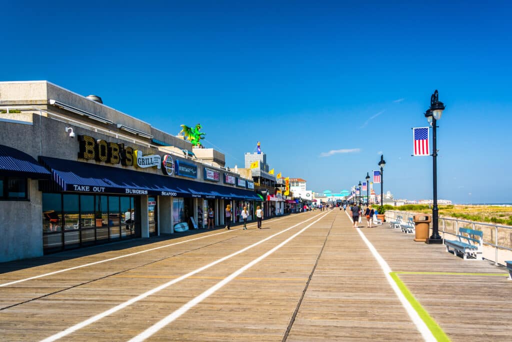 A sunny day on the Ocean City boardwalk features shops and eateries with awnings, including a place called Bob's Grill. People stroll along the wooden pathway, and a few benches and American flags are visible along the sides, reminiscent of the serene promenades near a drug and alcohol rehab treatment center.