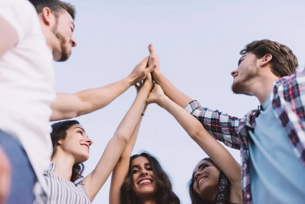 A group of five people standing in a circle with their arms raised, touching hands together in the center. They are smiling and appear to be celebrating or expressing unity, possibly as part of a drug rehab program, with a clear blue sky visible in the background.