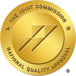 A gold circular emblem with the text "The Joint Commission" at the top and "National Quality Approval" at the bottom, encircling a geometric design of three stacked squares and diagonal lines, symbolizes excellence for drug and alcohol rehab treatment centers in New Jersey.